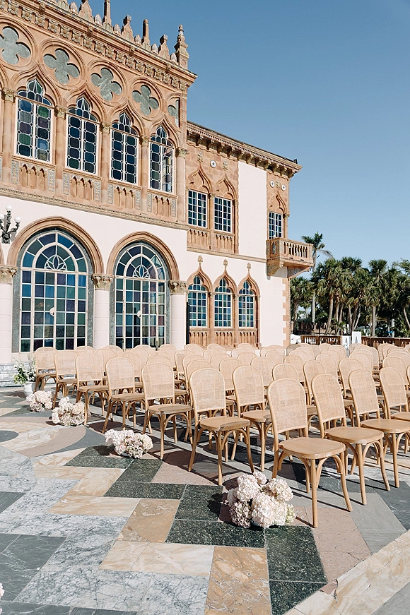 Outdoor ceremony setup with wedding ceremony chairs, light wood rows and white hydrangea aisle florals on a stone patio by arched windows and palms