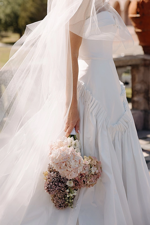 Bridal bouquet of hydrangea wedding bouquet blooms held against a strapless white dress with veil, in sunlight by a stone balustrade