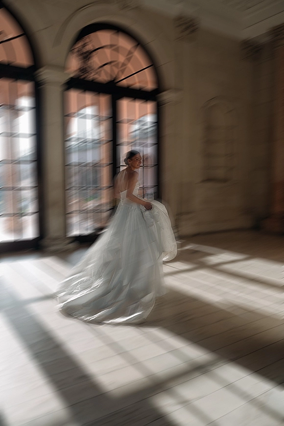 Bridal portrait of a bride twirling dress, holding a long train in window light by arched windows, stone walls, and tiled floor shadows