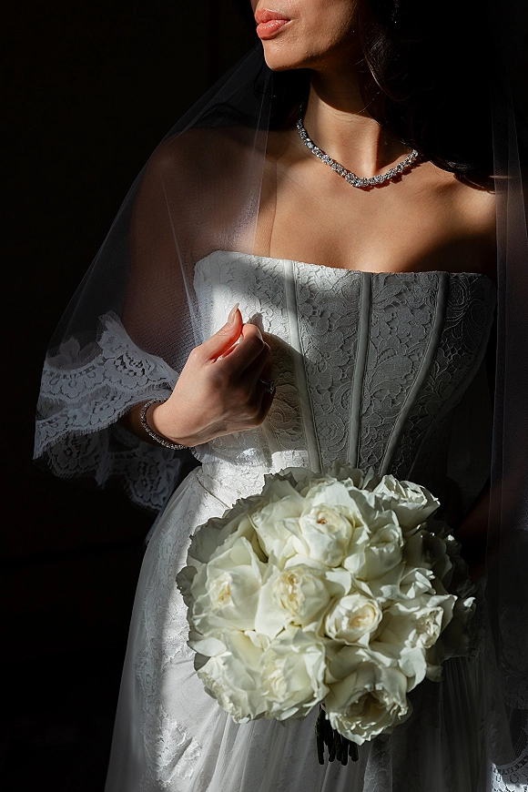 Bridal portrait of a bride in a strapless lace wedding dress holding a white rose bouquet, sheer veil and diamond necklace against a dark backdrop