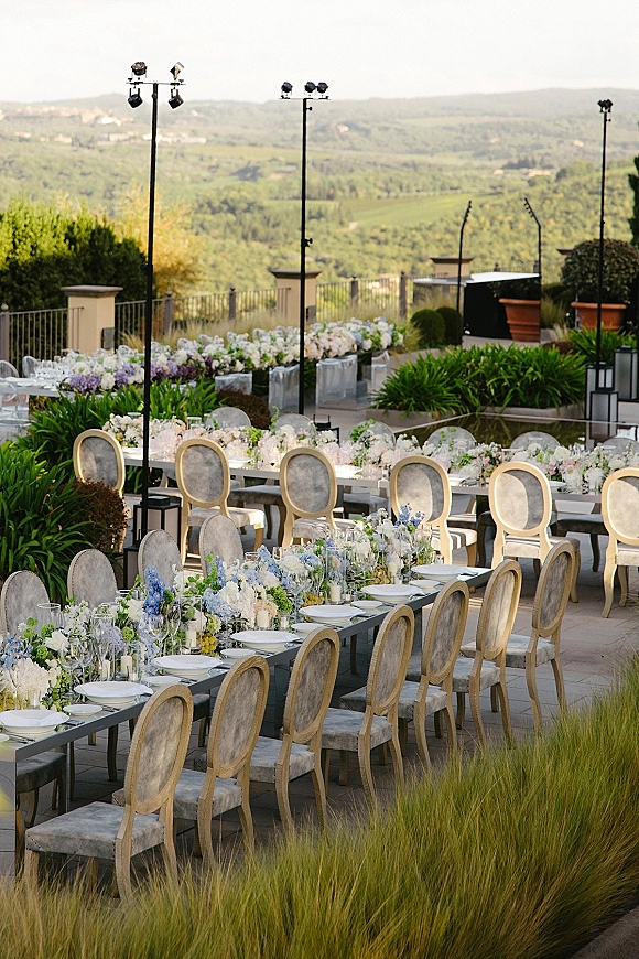Reception tablescape for an outdoor wedding reception with long banquet tables, white and blue florals, greenery garland, and candlelit place settings on a terrace patio overlooking rolling hills