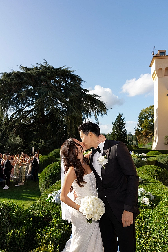 Wedding kiss portrait of bride and groom kissing as he dips her, veil flowing and white bouquet visible against garden hedges and villa sky
