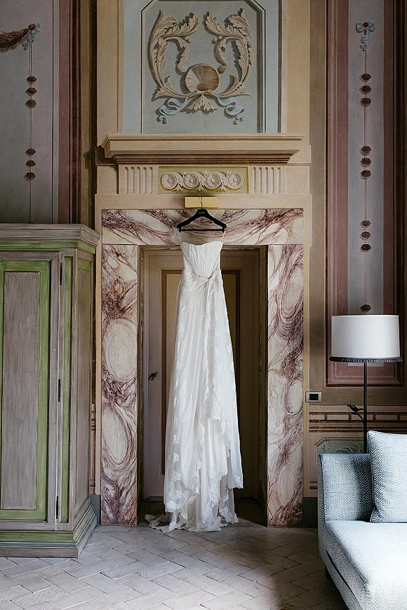 Wedding dress on hanger, strapless bridal gown with lace hem hanging by a marble fireplace in an ornate bridal suite interior