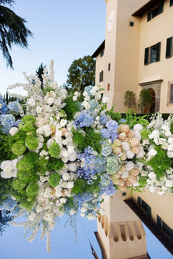 Wedding floral installation with hydrangea wedding flowers and roses on a reflective aisle, set before a villa exterior under blue sky