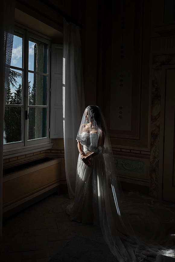 Bridal portrait of a bride in an off-the-shoulder lace gown with a long veil and train, standing by an open window with sheer curtains.