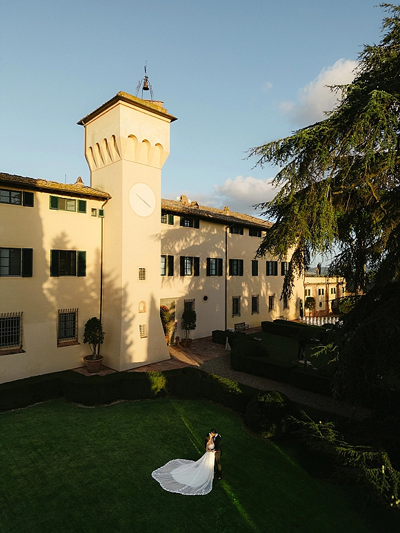 Couple portrait from above showing bride’s wedding dress train and veil beside groom in suit on a villa lawn near a clock tower