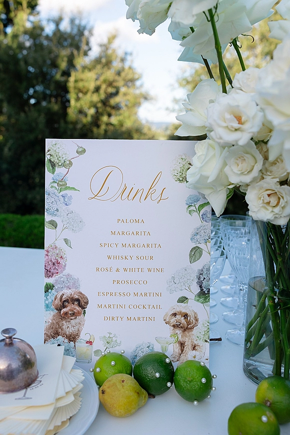 Wedding drink menu with signature cocktail menu sign beside white roses, wine glasses, limes and pear on a white outdoor tabletop with greenery