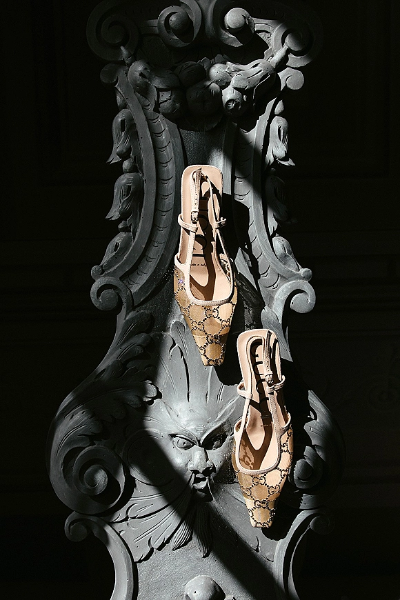 Bridal shoes in beige pointed-toe heels with ankle straps and monogram mesh fabric on an ornate black carved chair in dramatic shadow