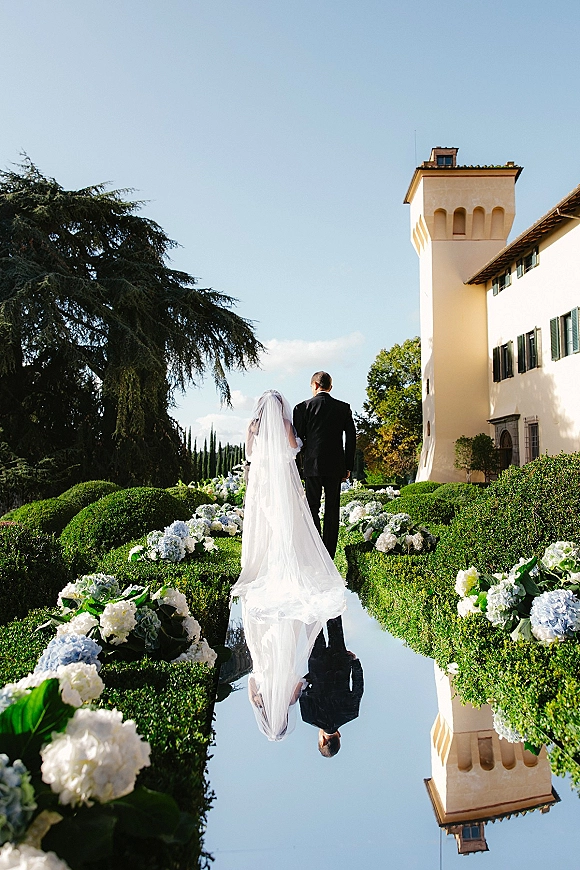 Wedding couple portrait of bride and groom walking away, her long veil and train flowing beside a reflecting pool at a villa garden estate