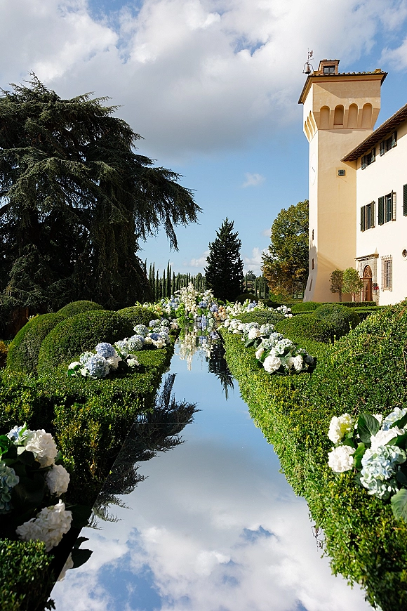Outdoor ceremony aisle with mirrored wedding aisle runner, lined with blue hydrangea flowers and hedges before a villa tower under clouds