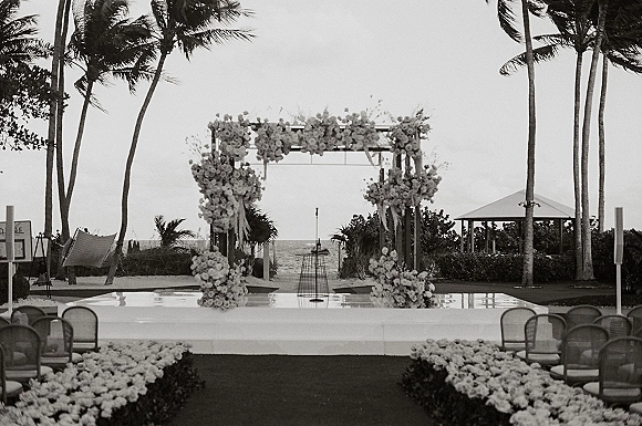Ceremony setup for a beach wedding ceremony with a floral arch framing the stage, aisle flowers, chairs, and ocean palms behind