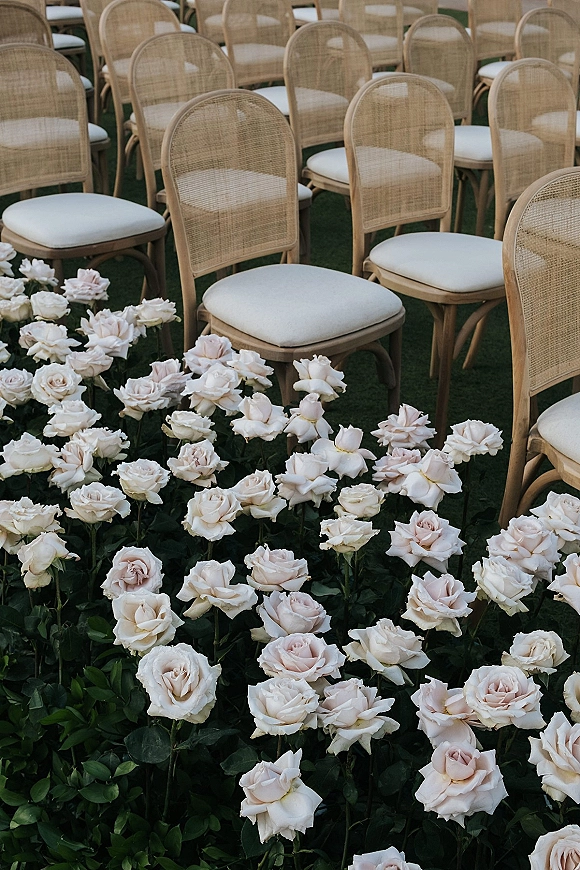Ceremony seating with outdoor wedding chairs featuring cane backs, white cushions, and rose greenery accents arranged on a grass lawn