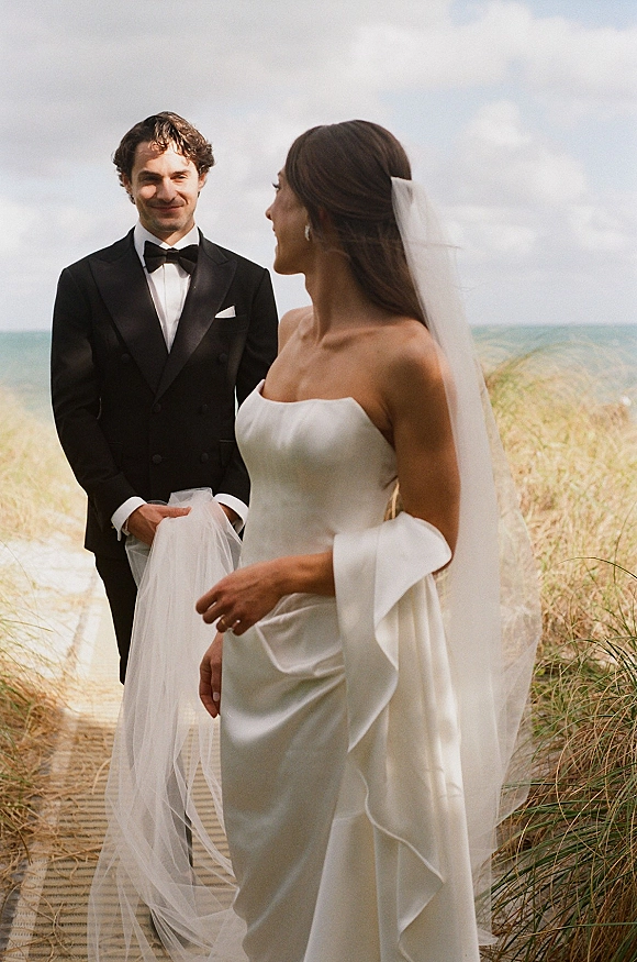 Couple portrait of bride looking at groom on a beach boardwalk, her veil blowing in dune grass beside the ocean under cloudy sky