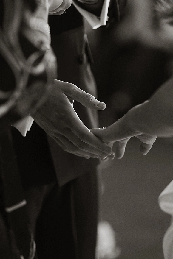 Ceremony handholding close-up of bride and groom holding hands, her sleeve and his suit and tie in focus against a softly blurred indoor backdrop