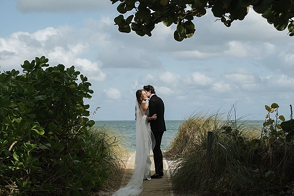 Wedding couple portrait of bride and groom hugging for a forehead kiss on a beach boardwalk, her long veil blowing by dunes and ocean