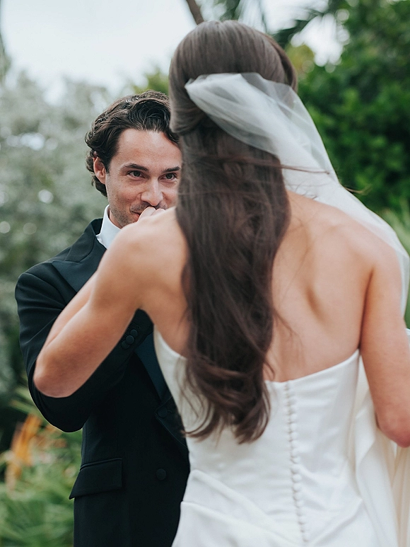 First look moment as bride in a strapless wedding dress and veil approaches groom in black tuxedo under palm trees and sky