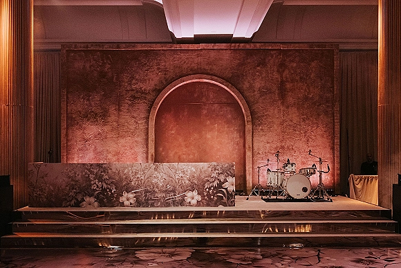 Wedding stage decor with a wedding band stage setup featuring an arched textured backdrop, floral front panel, drum kit, and warm uplighting in a ballroom