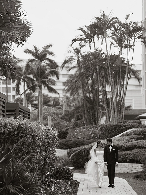 Couple portrait of bride and groom walking hand in hand, bride looking at groom as her veil blows along a palm-lined path near a hotel
