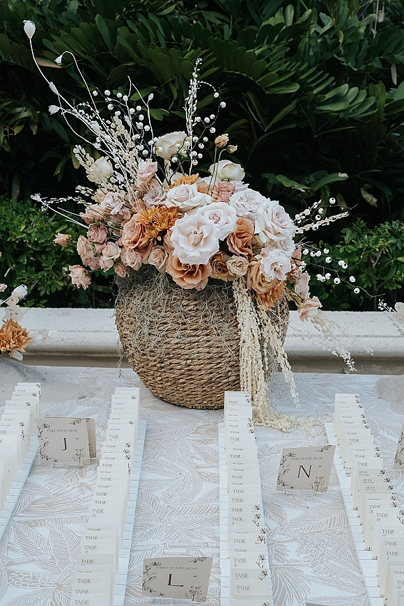 Wedding seating chart with an escort card display and roses, dried florals, and hanging amaranthus on white linens beside green foliage and stone ledge