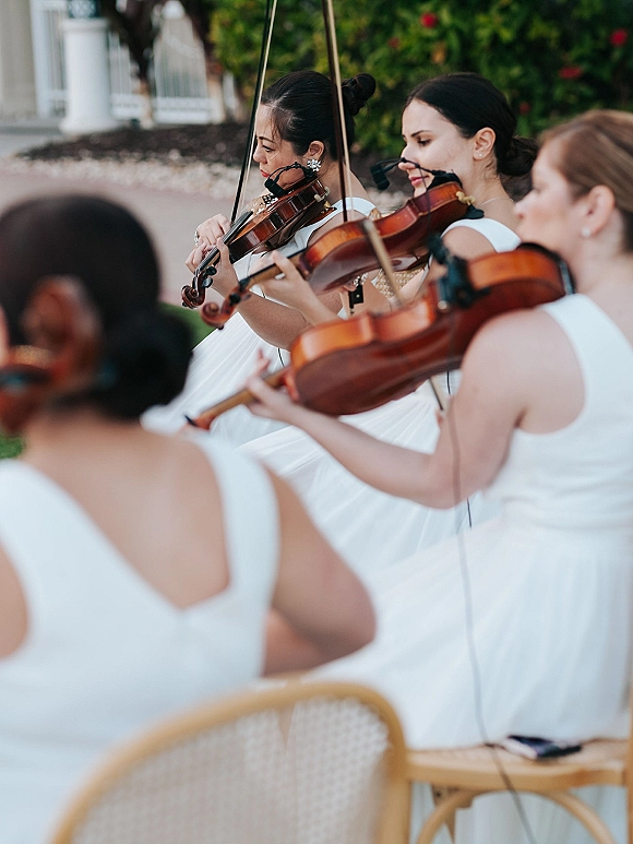 Wedding musicians in white dresses playing violins with bows along a garden walkway with greenery and white railing for ceremony music
