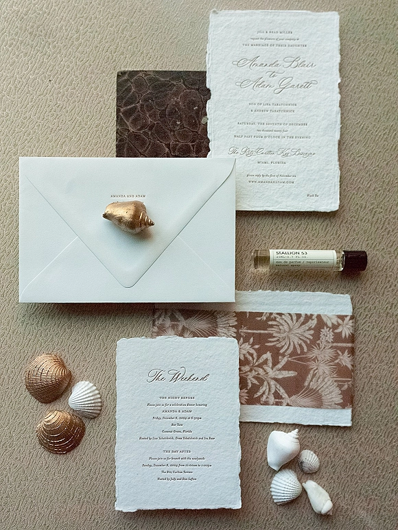 Invitation suite with deckle edge wedding invitations, calligraphy cards, envelopes, seashells, and a perfume vial on beige fabric backdrop
