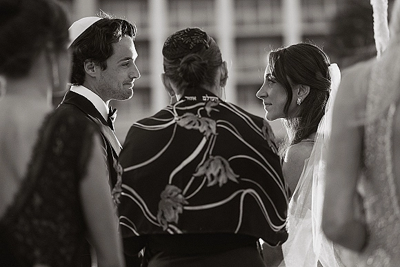Wedding ceremony moment at a Jewish wedding ceremony, bride in veil and drop earrings faces groom in tuxedo as officiant stands between them outdoors