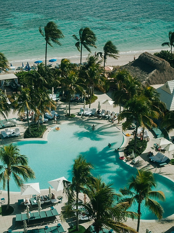 Beach resort pool with palm trees and loungers beside turquoise water, with sun umbrellas and an ocean shoreline beyond