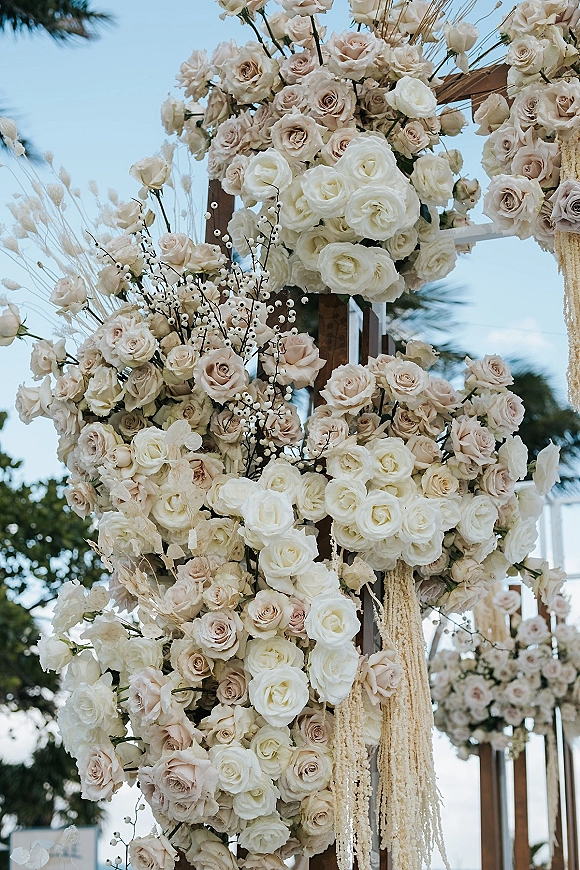 Wedding floral arch with blush and ivory roses, dried grasses, and hanging garland on a wooden frame against blue sky and trees