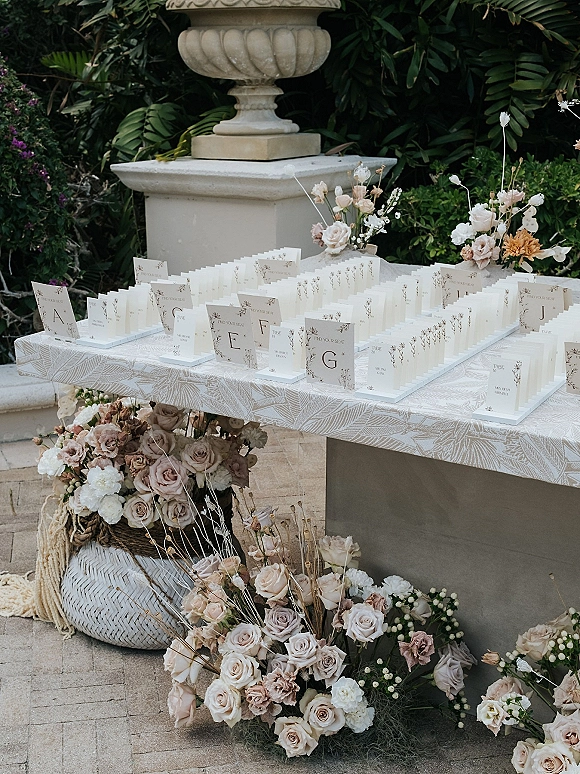 Wedding escort cards in an escort card display with table numbers, set on a patterned cloth beside lush florals on a stone patio in a garden