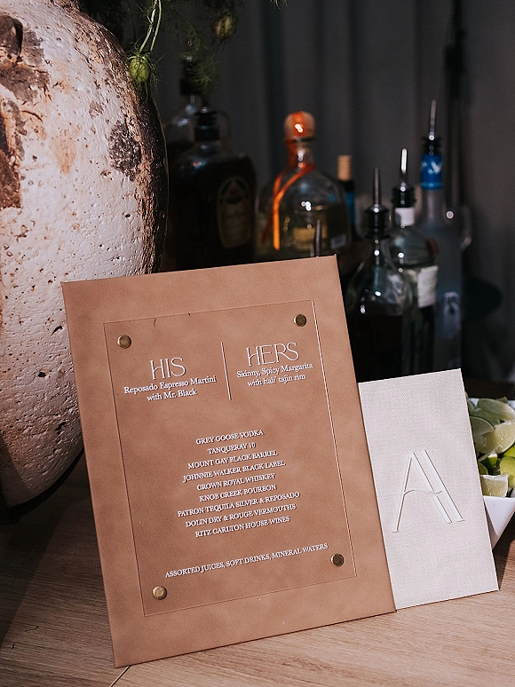 Wedding bar menu card with brass rivets listing signature cocktails beside liquor bottles, limes, and monogram napkins on a wood tabletop