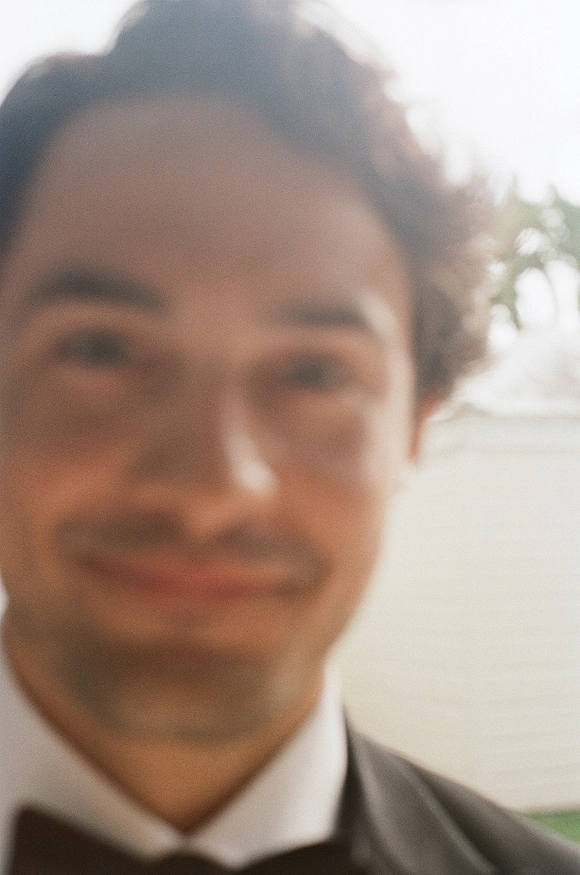 Groom portrait in a black tuxedo and bow tie with white dress shirt, standing outdoors against greenery and bright sky