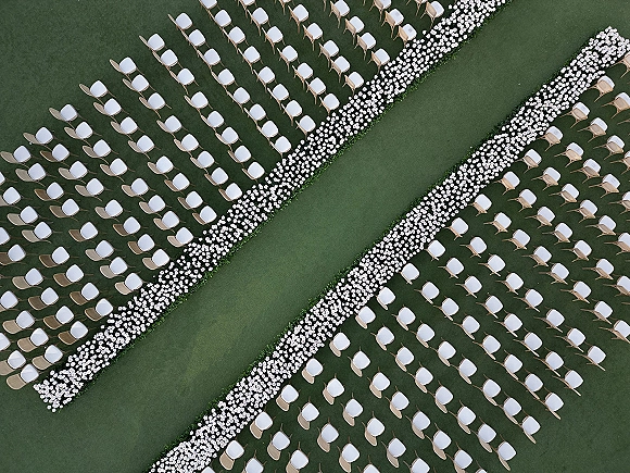 Ceremony aisle design with outdoor wedding aisle of white flowers and greenery border on grass, flanked by rows of white chairs with wooden frames
