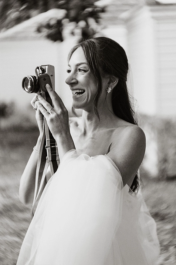 Bride portrait holding camera with a pearl bracelet, strapless tulle wedding dress, and veil, laughing in outdoor greenery backdrop