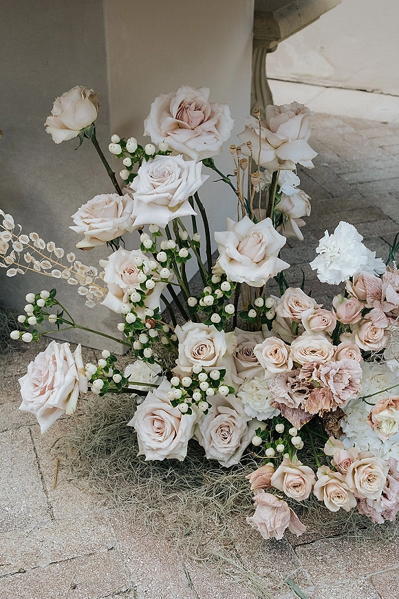 Wedding floral arrangement of blush and white roses with carnations and berries, grounded on Spanish moss beside a stone column floor