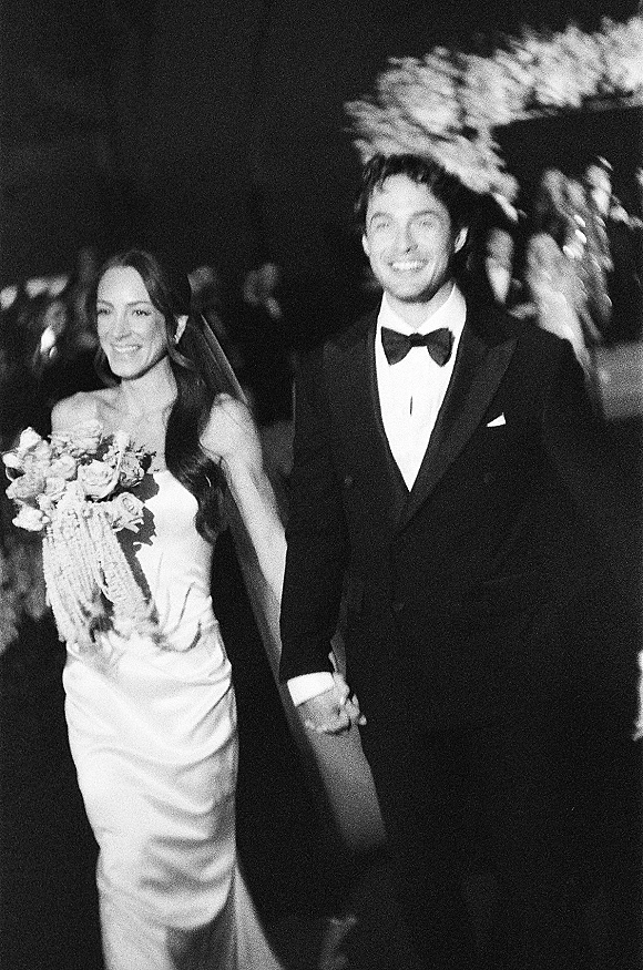 Wedding recessional as bride and groom walking hand in hand at night, her veil and bouquet visible while guests line a greenery walkway