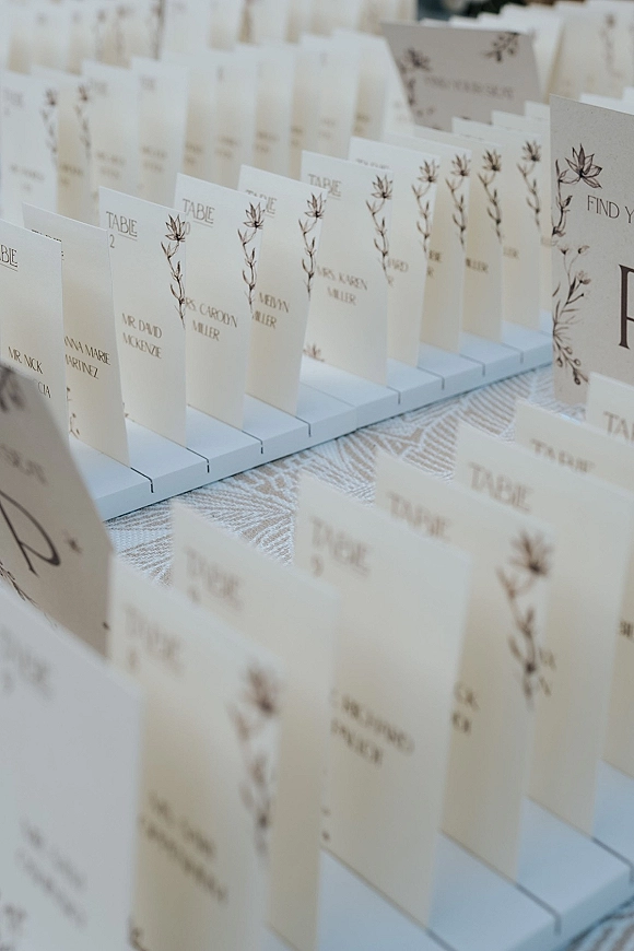 Wedding escort cards in an escort card display, neatly arranged on holders with calligraphy and botanical line art on a linen tablecloth
