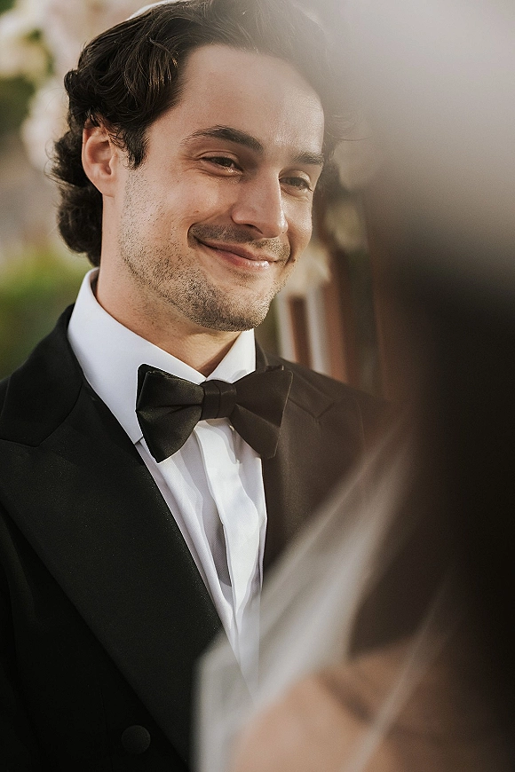 Groom portrait in a black tuxedo with bow tie, looking toward a blurred bride in the foreground against soft outdoor greenery