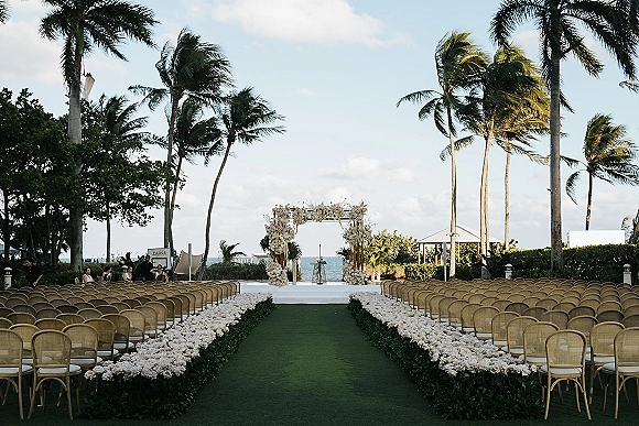 Ceremony setup for an outdoor wedding ceremony with a white floral arch, wicker chairs, and flower-lined aisle facing the ocean view