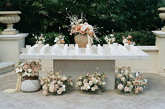 Escort card table with wedding escort cards arranged alphabetically on white linen, framed by blush roses and baskets on a stone patio backdrop