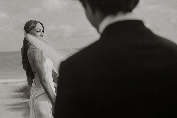 Wedding couple portrait on the shoreline as the bride looks back holding her veil, satin strapless dress flowing, ocean and sky behind