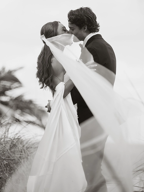 Wedding kiss portrait of bride and groom kissing beneath her veil, strapless dress and tuxedo with bow tie in beach dunes and tall grass