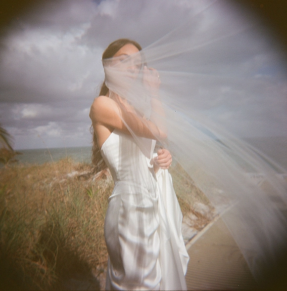 Bridal portrait of a bride holding veil in a strapless wedding dress on a cloudy beach, windswept beside dunes and boardwalk