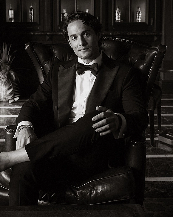 Groom portrait in a black tuxedo with bow tie, seated in a leather armchair in a dark wood lounge with liquor bottles behind him
