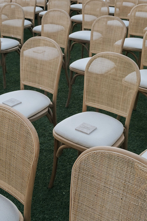 Ceremony seating with outdoor ceremony chairs, cane back chairs and white cushions lined in rows on a grass lawn with programs placed on seats