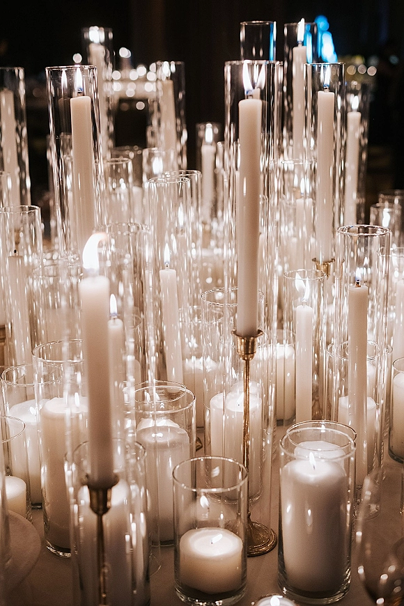 Wedding candle decor with glass hurricane vases and white taper and pillar candles in gold holders on a tabletop in dim reception lighting
