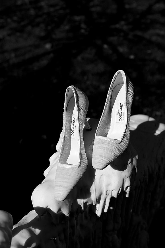 Wedding shoes with pleated high heel pumps perched on a stone statue, framed by dark foliage for an elegant bridal detail shot