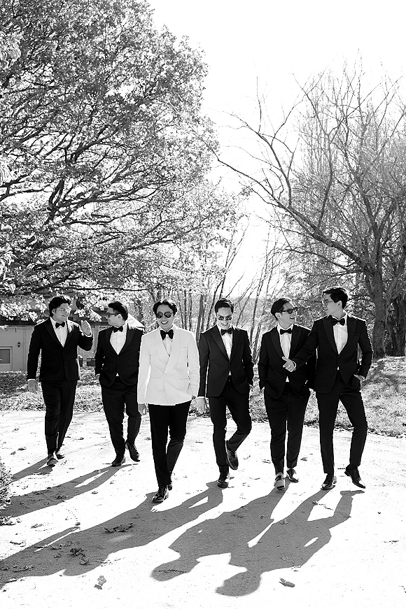 Groomsmen portrait of the groom in a white dinner jacket walking with tuxedoed friends in sunglasses on a sunlit tree-lined path