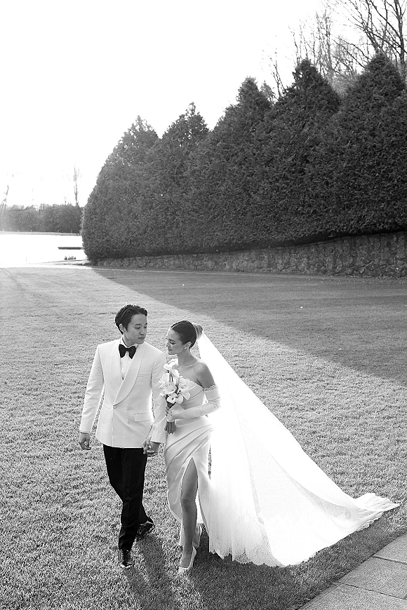 Couple portrait of bride and groom walking hand in hand, her veil flowing as she carries a calla lily bouquet on a lawn path