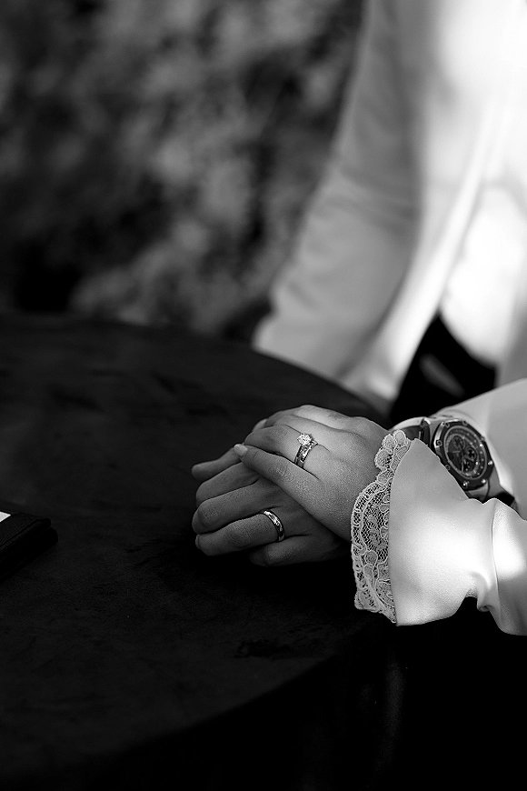 Wedding rings and engagement ring close up on clasped hands, bride’s lace cuff and groom’s wristwatch visible on an upholstered sofa indoors