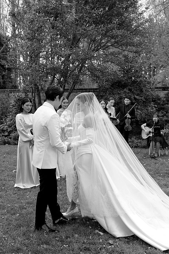 Wedding vows during an outdoor wedding ceremony as bride and groom hold hands, her cathedral veil trailing on a garden lawn by a stone wall